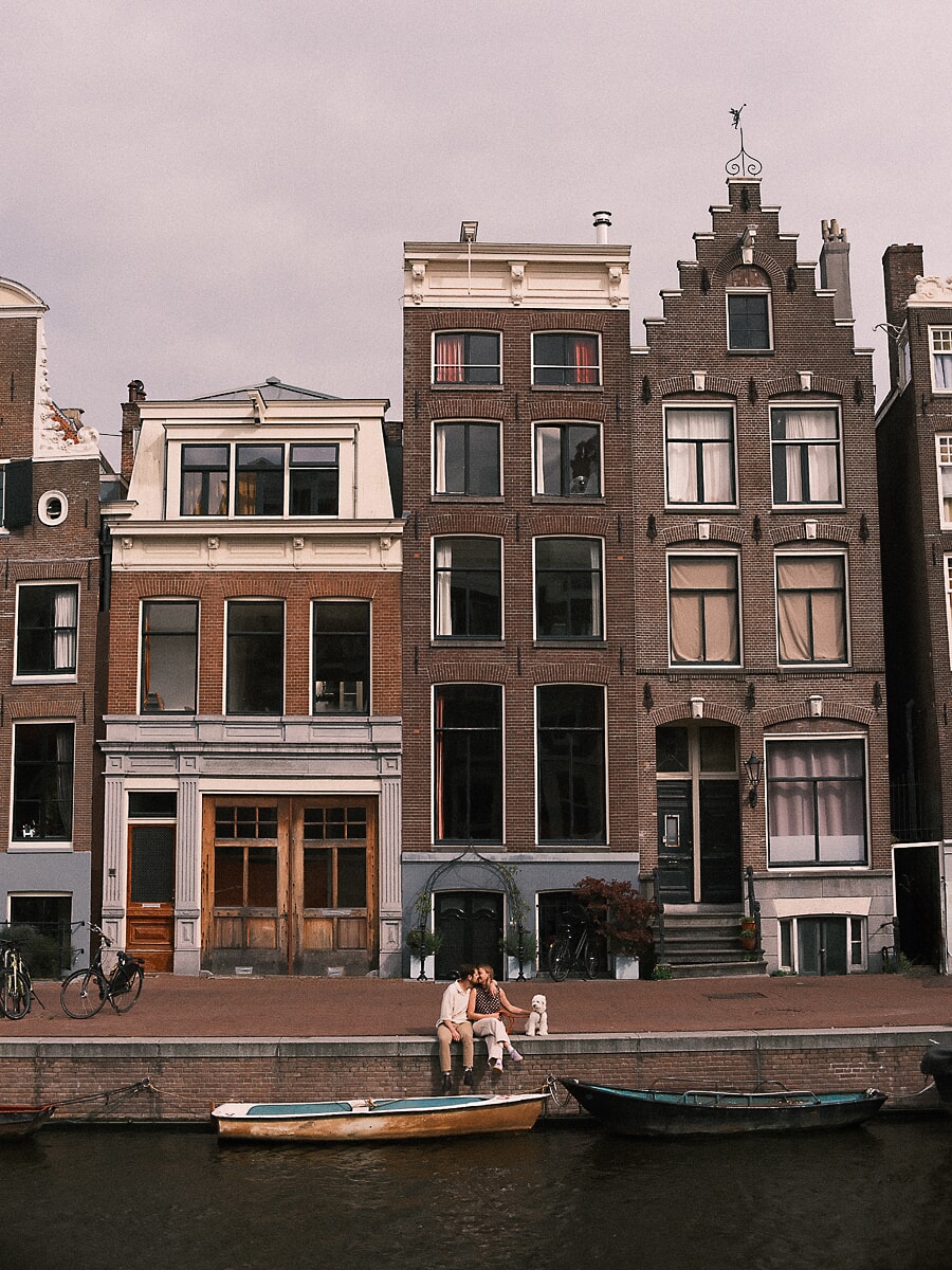 Engagement SP5 Wide-frame couple photography in Amsterdam capturing partners kissing on a canal edge in front of gabled houses