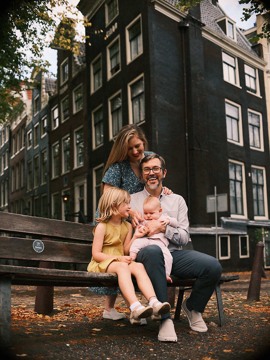 Family SP10 Family of four on a wooden bench among fallen leaves by a canal, the eldest girl in yellow beside her baby sibling, captured through cinematic family photography