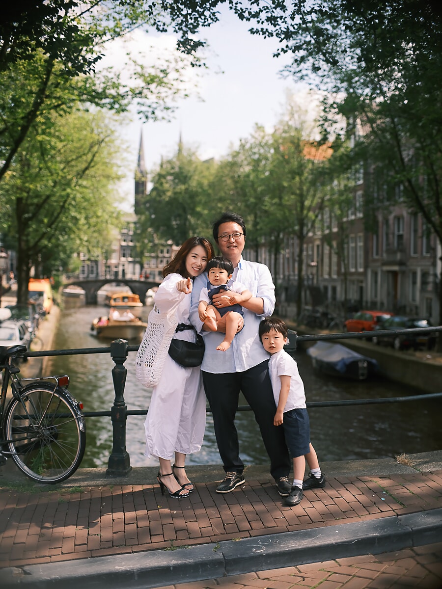 Family SP12 Expat family on a canal bridge in summer during a family photoshoot in Amsterdam, the parents holding a toddler beside their young son