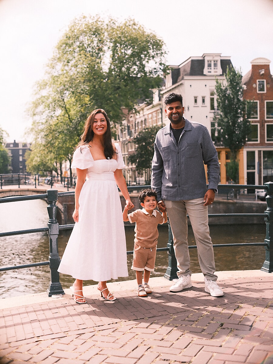 Family SP7 Smiling parents holding their young son's hands on a sunlit Amsterdam bridge, the mother in a white dress during a family shoot in summer