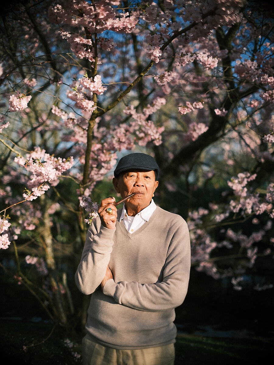 Group Family SP2 Expressive portrait of a grandfather in a flat cap pausing beneath blossoming cherry branches, part of a multigenerational family shoot