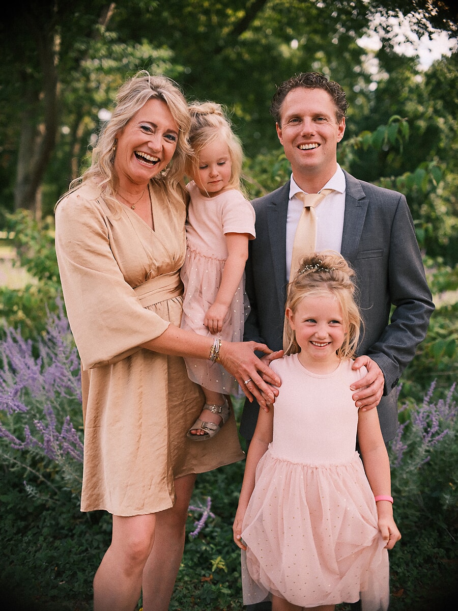 Group Family SP3 Parents and their two daughters laughing together among lavender in a green garden, the girls in soft pink dresses, a warm moment of family photography in Amsterdam