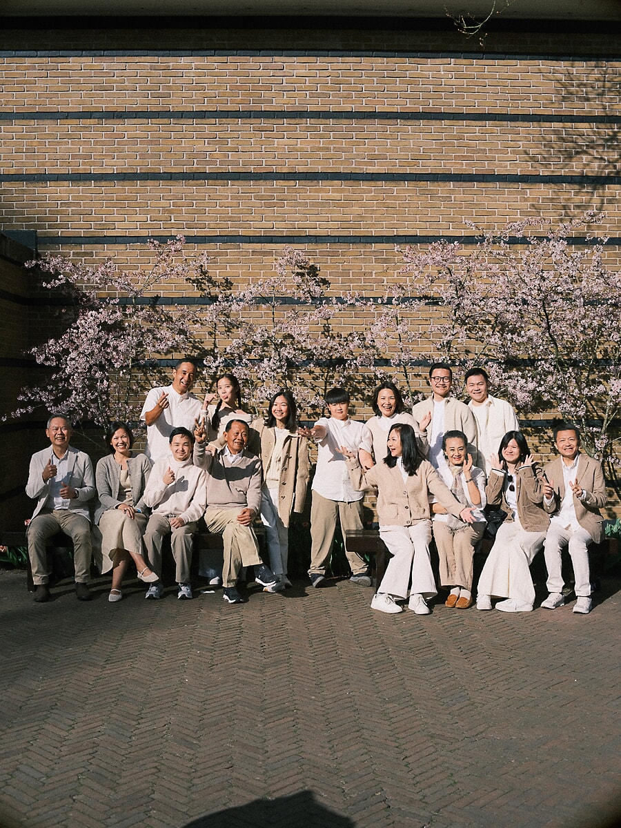 Group Family SP8 Large family multigenerational photoshoot in cream tones, relatives of all ages gathered before a brick wall and blossoming cherry tree