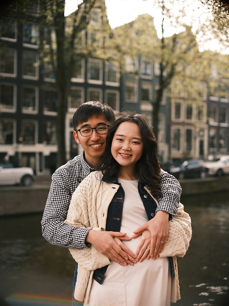 Maternity SP1 Smiling couple beside an Amsterdam canal as the partner wraps his arms around the mother-to-be to shape a heart over her pregnant belly during their pregnancy photoshoot