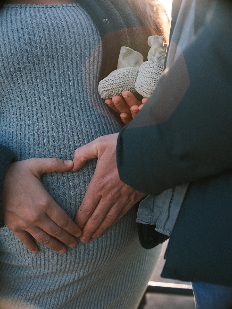 Maternity SP3 Backlit bump photoshoot showing a couple's hands holding tiny knitted baby shoes against a pregnant belly in a blue dress
