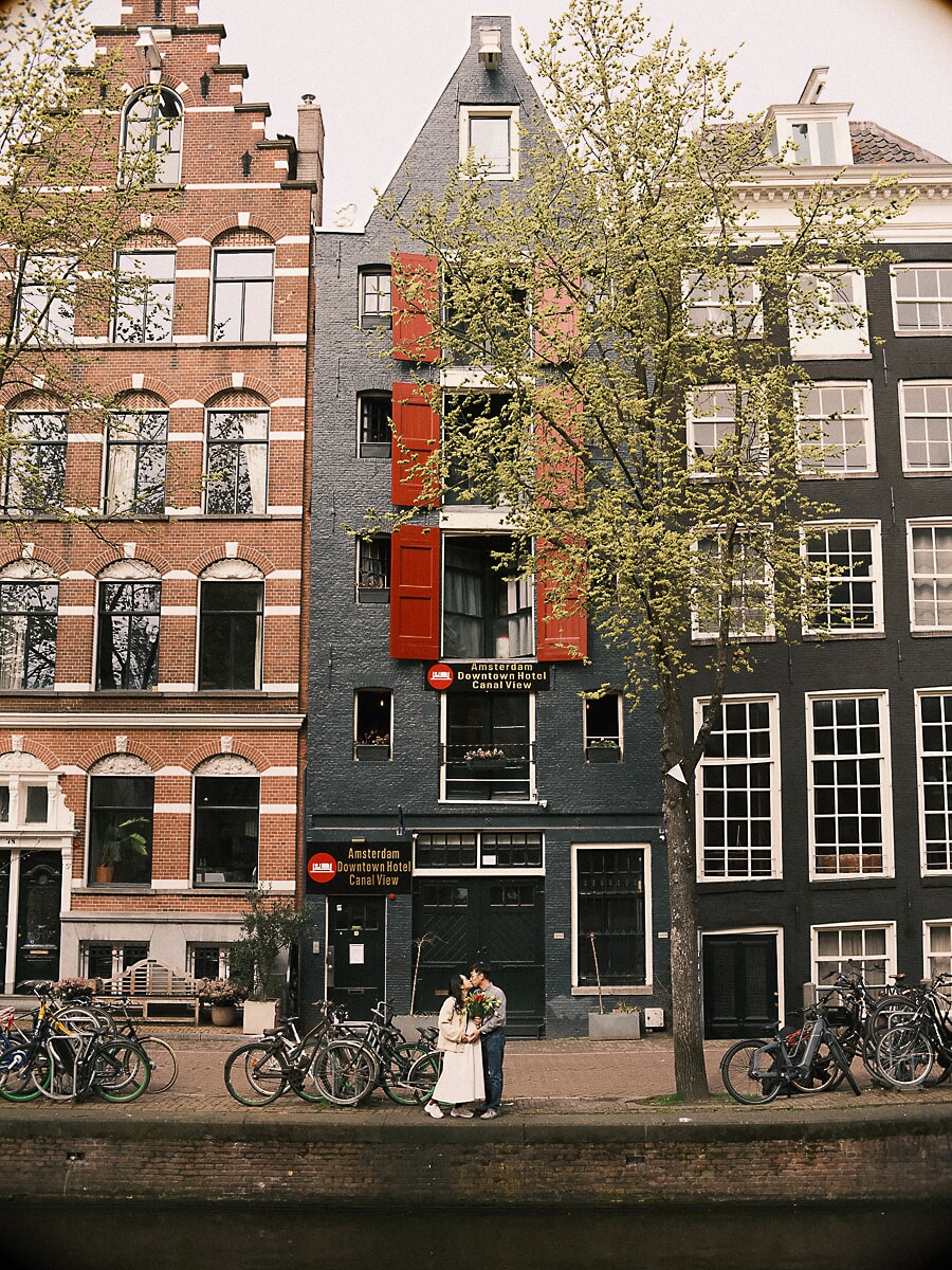 Maternity SP4 Tall canal houses with red shutters rising above a couple kissing among parked bicycles during their maternity photoshoot in Amsterdam