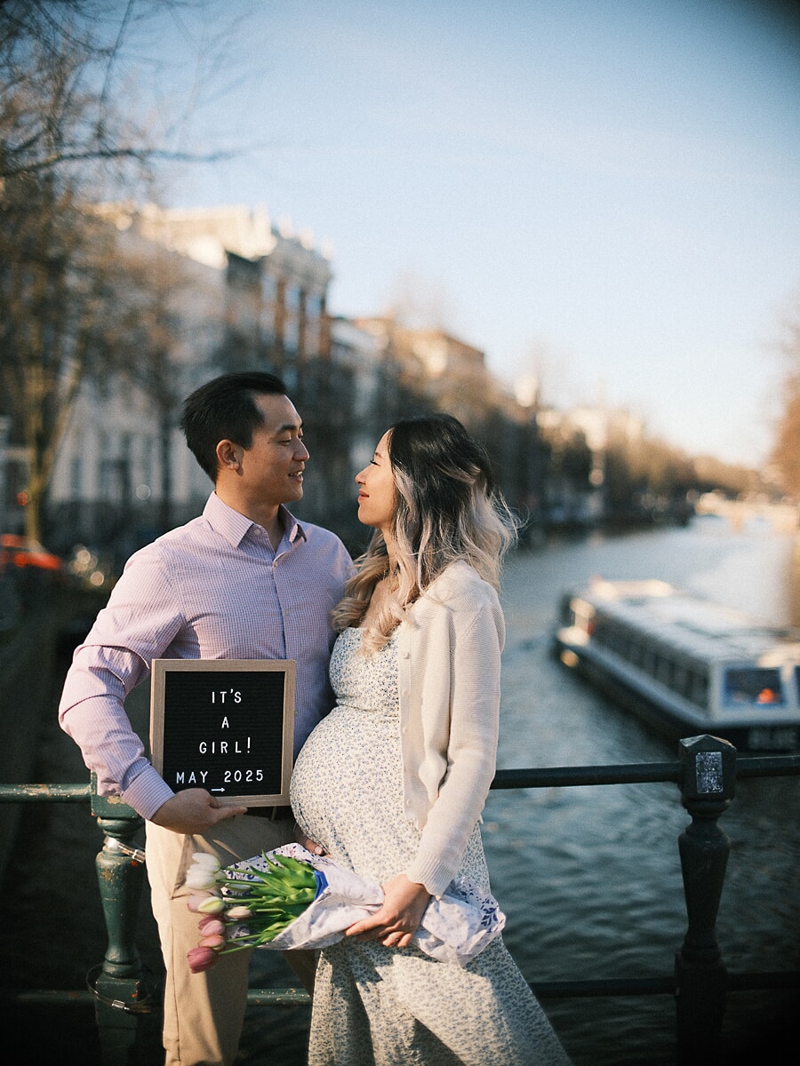 Maternity SP6 Gender reveal photoshoot by an Amsterdam canal as a couple holds an "It's a girl" letter board and a tulip bouquet, a tour boat drifting behind them