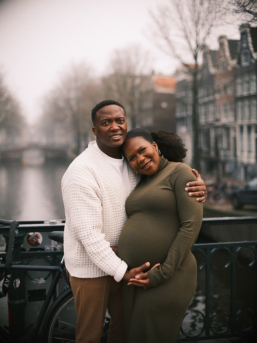 Maternity SP7 Joyful maternity portraits of a couple cradling the pregnant belly beside a canal railing, the expectant mother in an olive dress and her partner in a cream knit sweater