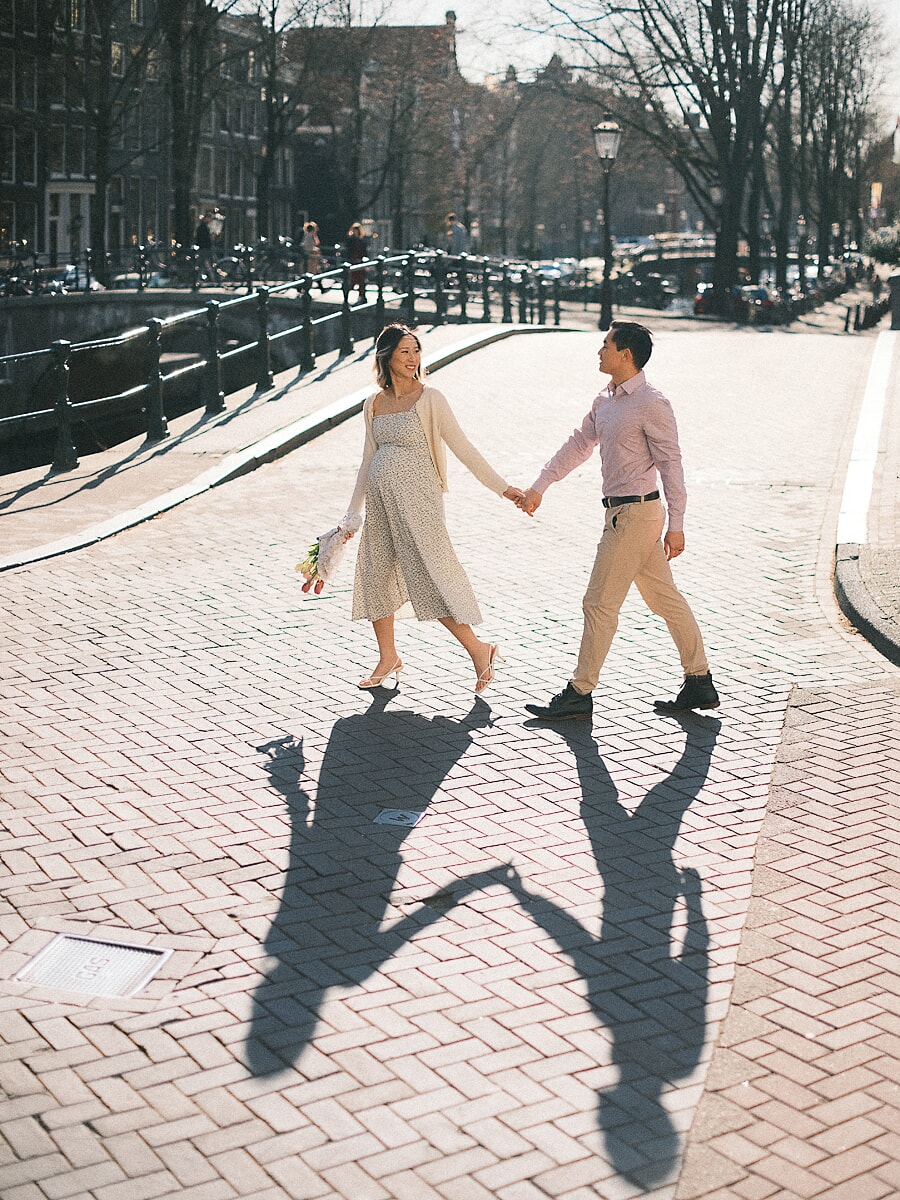 Maternity SP8 Pregnant woman and her partner walking hand in hand across a sunlit Amsterdam bridge during a babymoon photoshoot, long shadows trailing over the cobblestones
