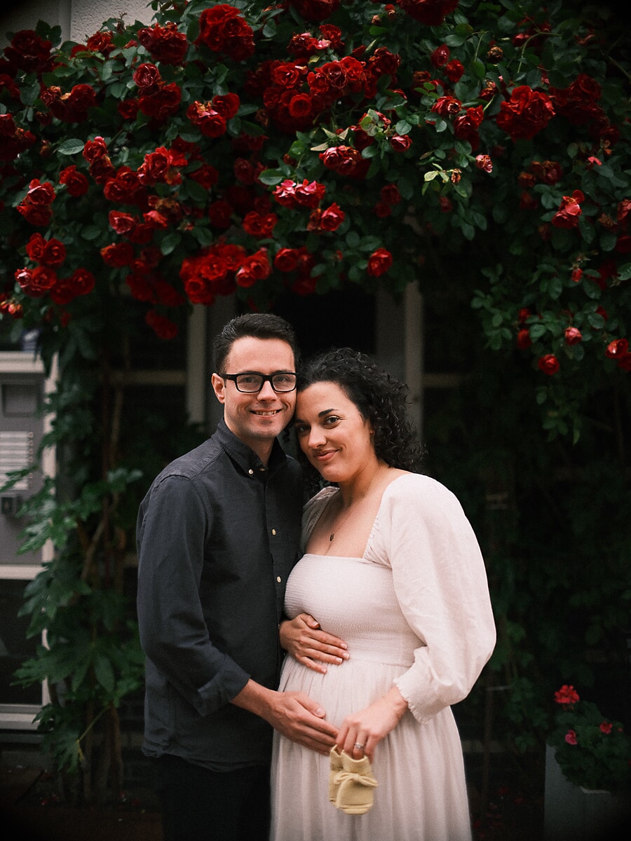Maternity SP9 Expecting parents in a maternity session beneath a wall of red climbing roses, holding small knit baby booties against the mother's belly