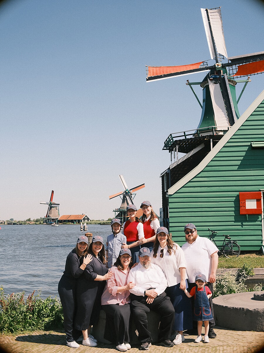 Group Family SP12 Large multigenerational family in matching caps gathered by the water at the Zaanse Schans windmills, three generations together for a family reunion photoshoot on a clear summer day
