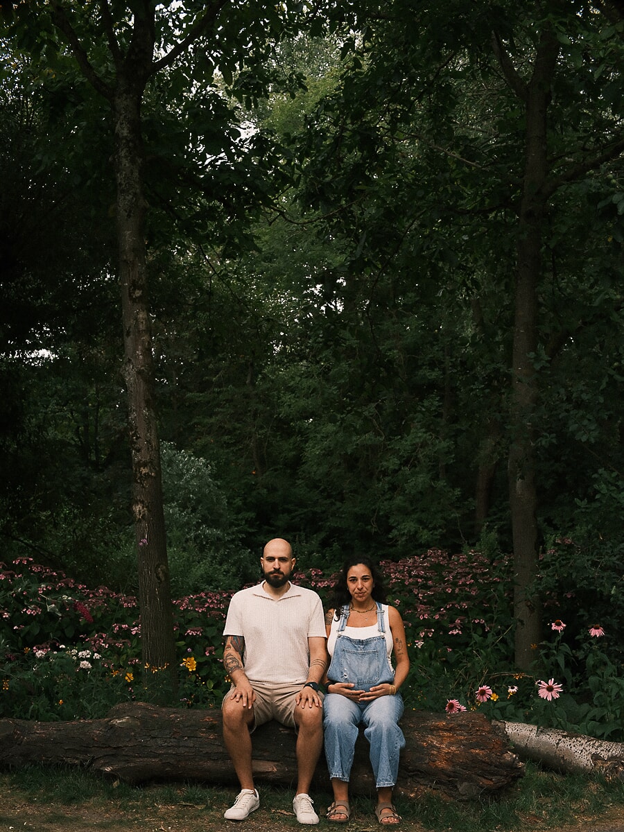 Maternity SP10 Relaxed pregnancy photoshoot of a couple seated on a fallen log in a lush flower garden, the expectant mother in denim dungarees resting a hand on her baby bump