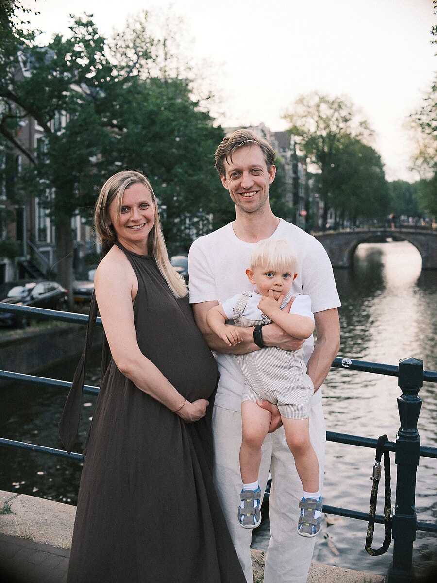 Maternity SP11 Growing family on a sunlit canal bridge during a summer maternity session, the father cradling their young son beside the mother-to-be in a dark sleeveless dress, an arched bridge behind them