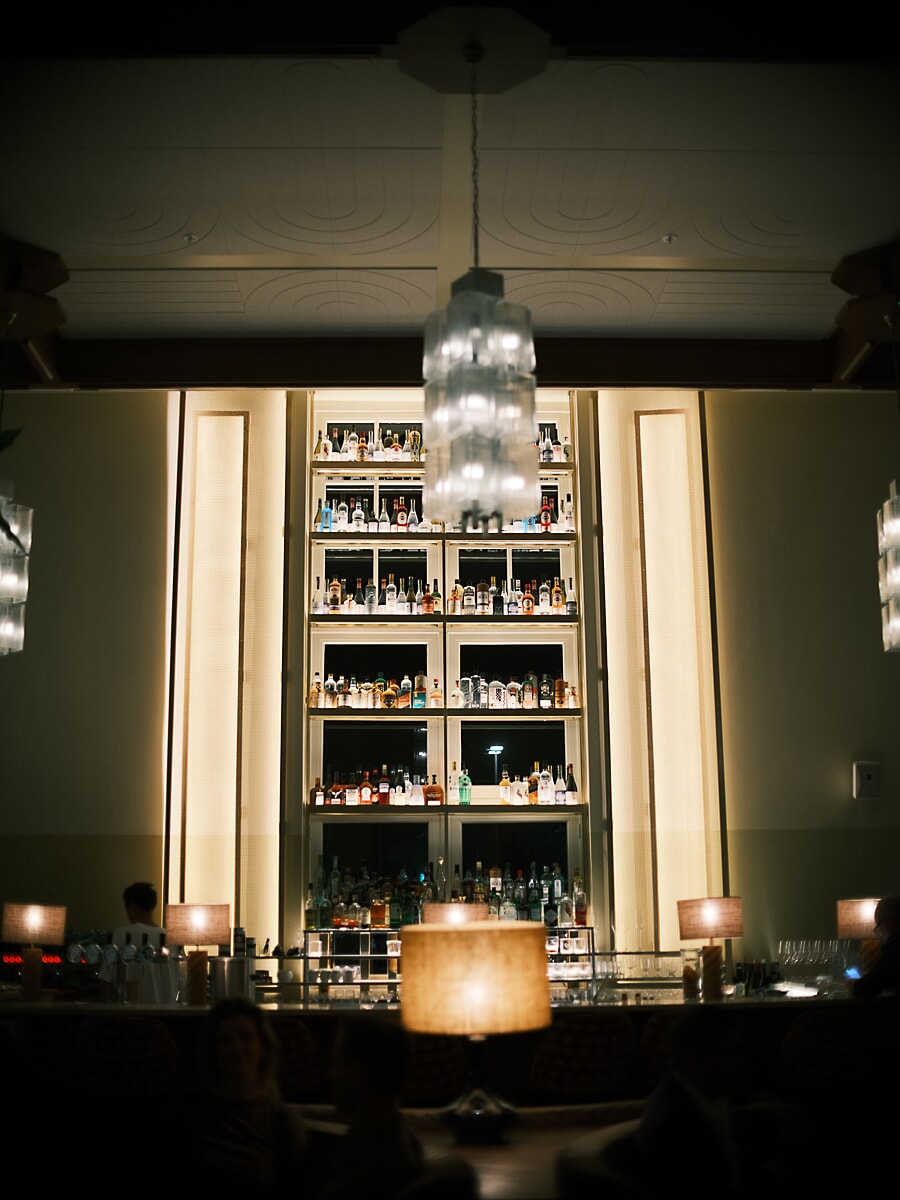 Art deco bar seen head-on, tall lit bottle shelves and a fluted chandelier above the table lamps