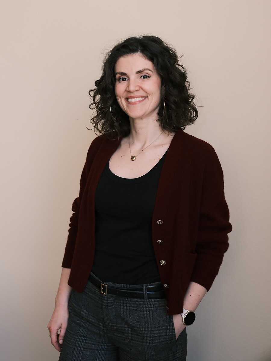 Woman with curly hair in a burgundy cardigan and checked trousers, one hand in her pocket against a beige wall