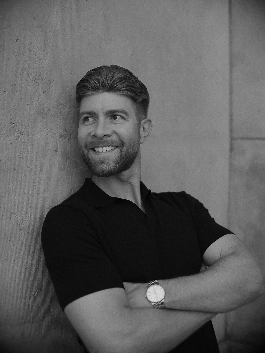 Man in a black polo with his arms folded against a concrete wall, looking off with a broad smile, in black and white