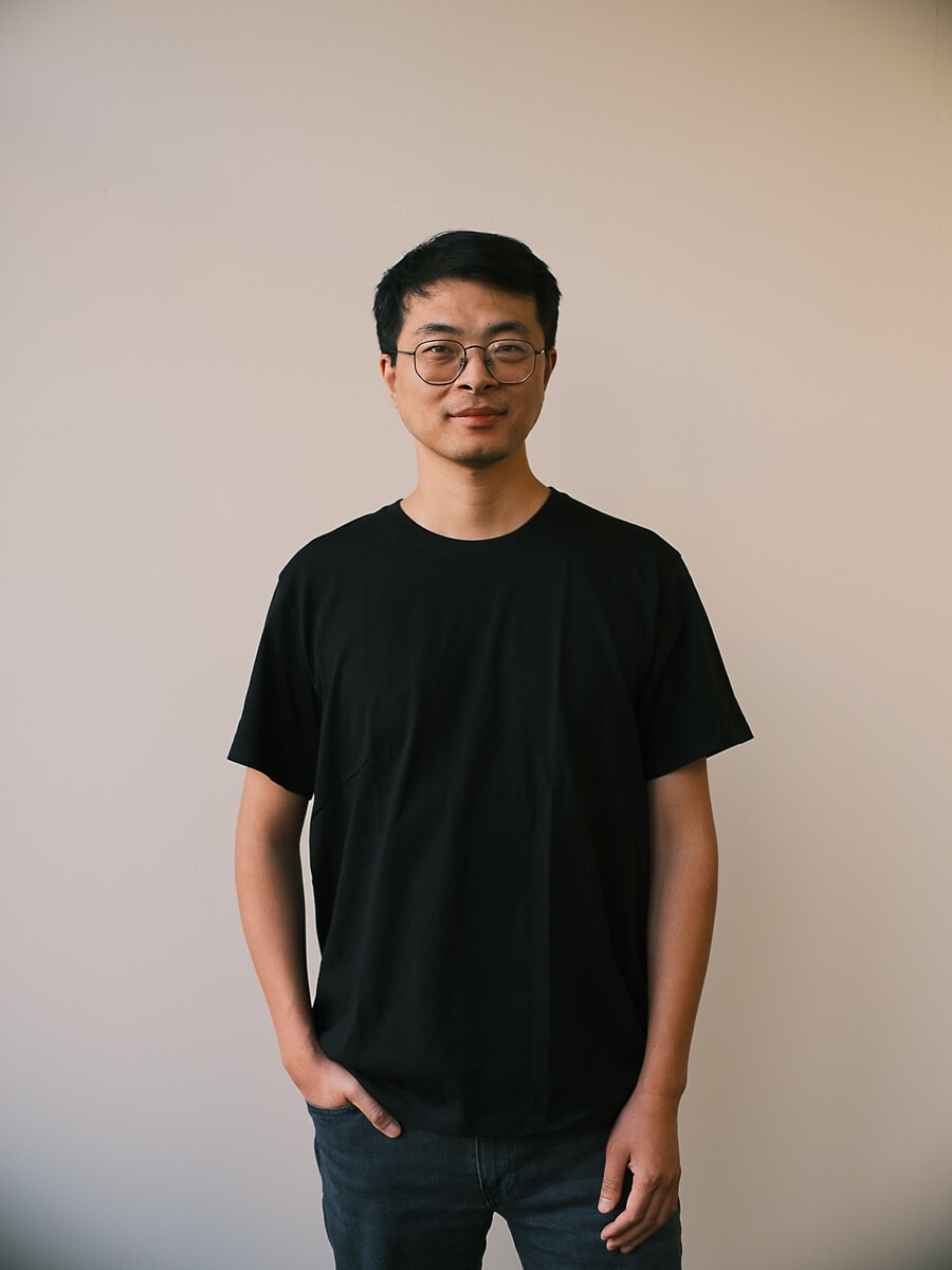 Man in round glasses and a black tee with his hands in his pockets against a plain pale wall, a corporate portrait in Amsterdam