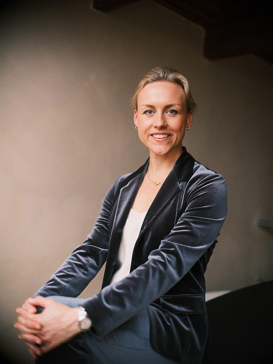 Woman in a grey velvet blazer perched on a stool, plaster wall and timber beams behind, a business portrait session in Amsterdam