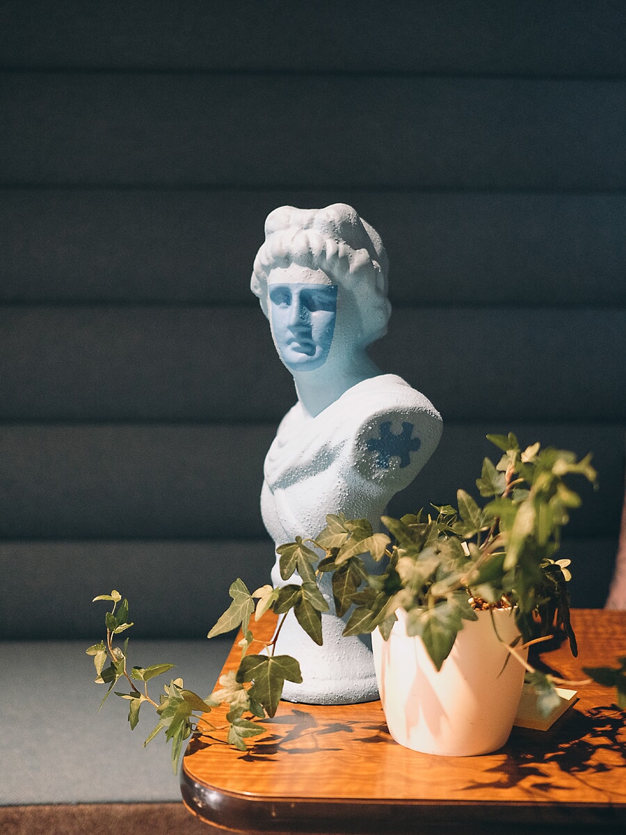 Pale blue classical bust and a pot of ivy on a wooden table against a dark ribbed banquette, sunlight falling across them