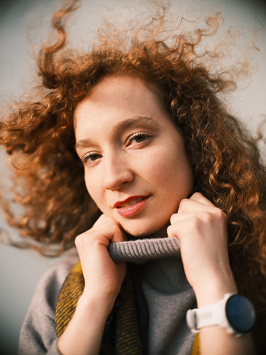 Very close portrait of a woman with red curls holding a grey rollneck up to her chin against an open sky