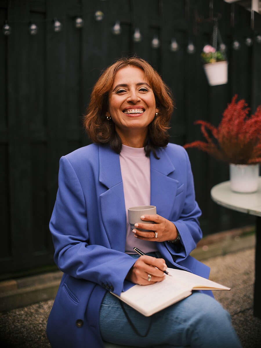 Coach in a blue blazer laughing with a coffee cup and open notebook under festoon lights, a branding photoshoot in Amsterdam