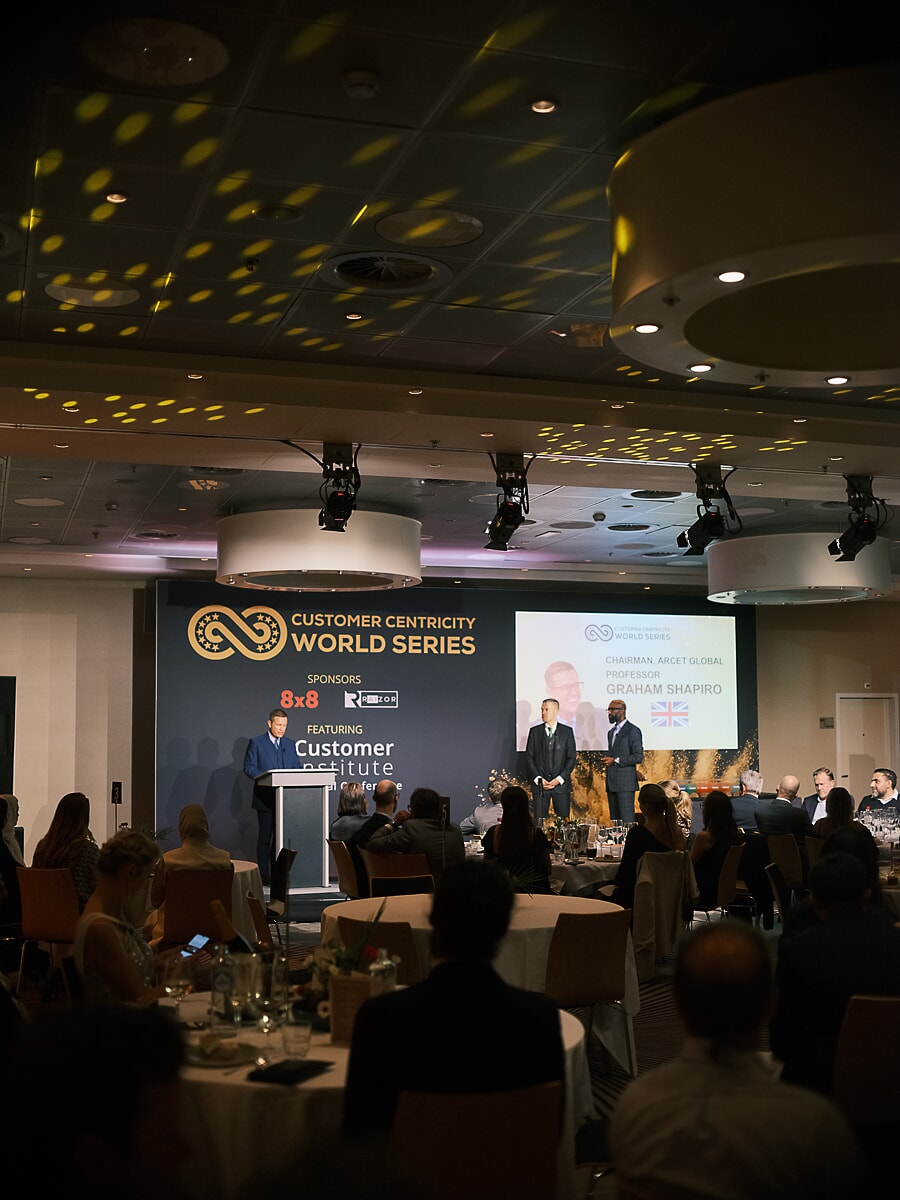Wide view of a conference dinner, the speaker at his lectern and a portrait slide above the seated audience