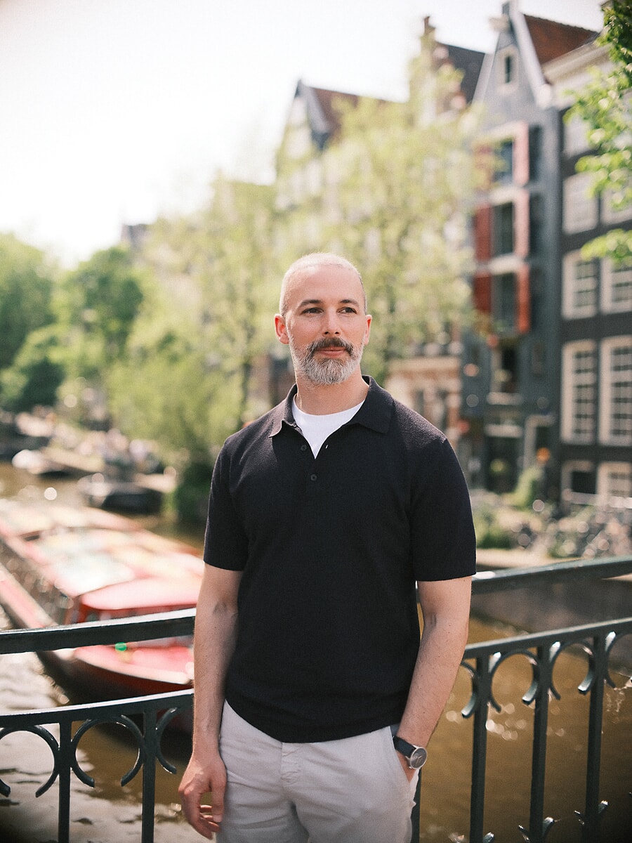 Man with a grey beard in a navy polo standing at a canal railing with moored boats and gabled houses behind, a confidence portrait in Amsterdam