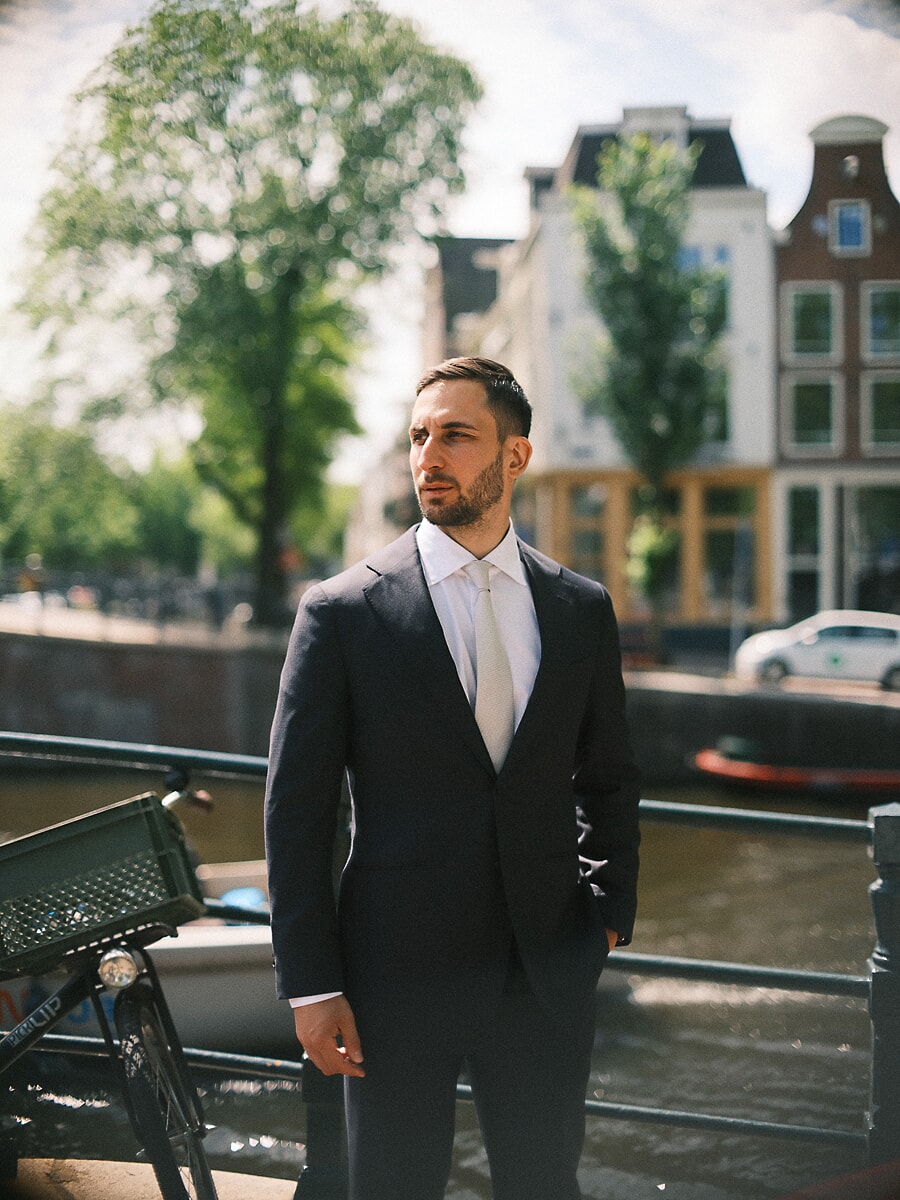 Man in a navy suit and tie looking off past a canal railing, bright gabled houses behind him, a formal confidence portrait in Amsterdam