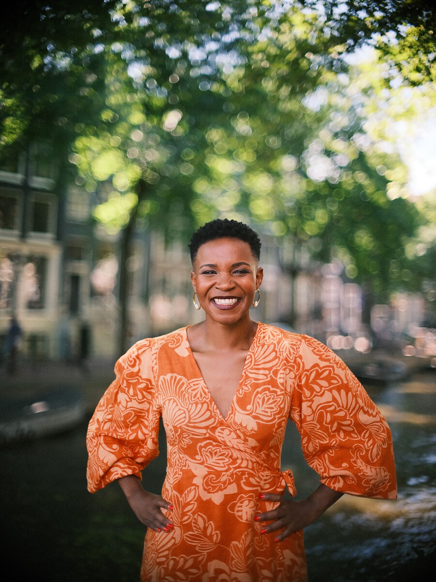Woman in an orange leaf-print dress laughing with her hands on her hips, dappled canal light behind her, a lively confidence portrait