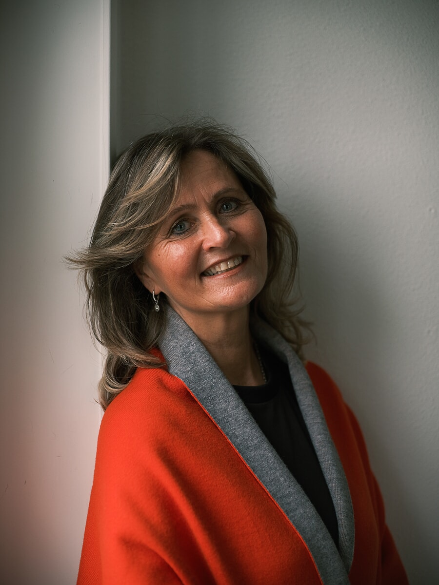 Woman with layered blonde hair smiling in a red and grey wrap coat against a plain pale corner, an indoor portrait from a confidence photoshoot