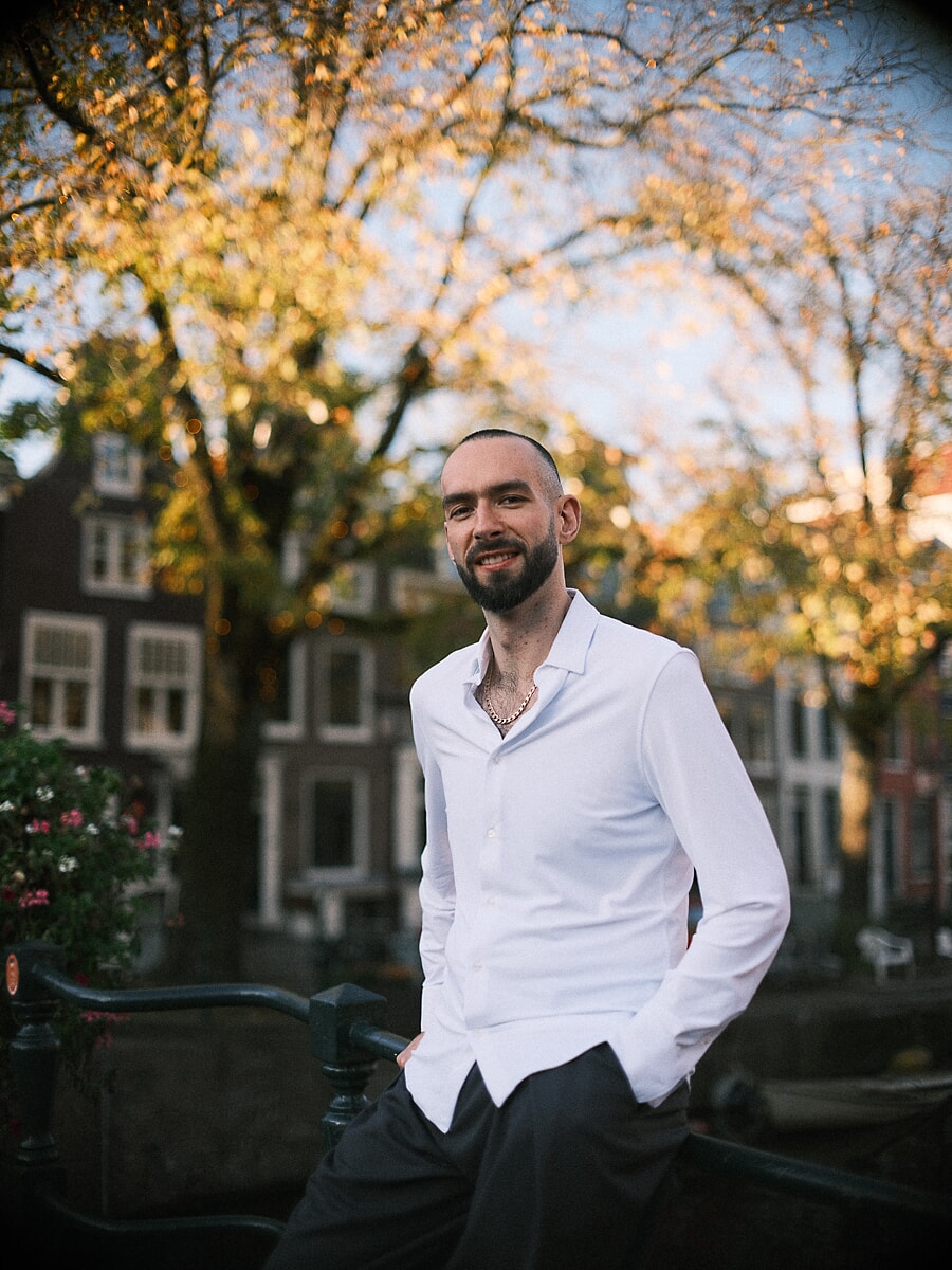Man in a white shirt leaning on a canal railing beneath golden autumn leaves, a warm confidence portrait in Amsterdam