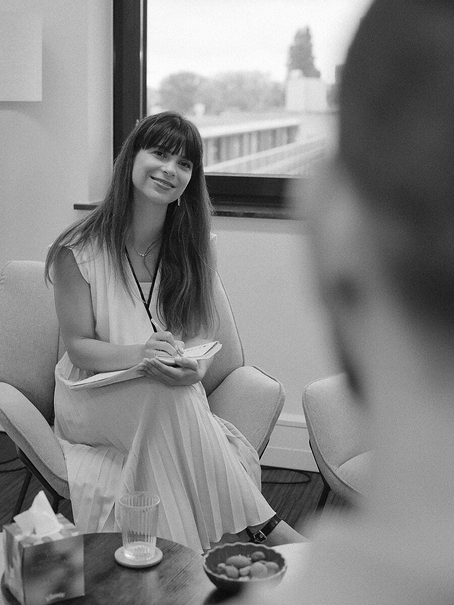 Consultant in a pleated dress taking notes in an armchair while she listens, a colleague blurred in the foreground, in black and white
