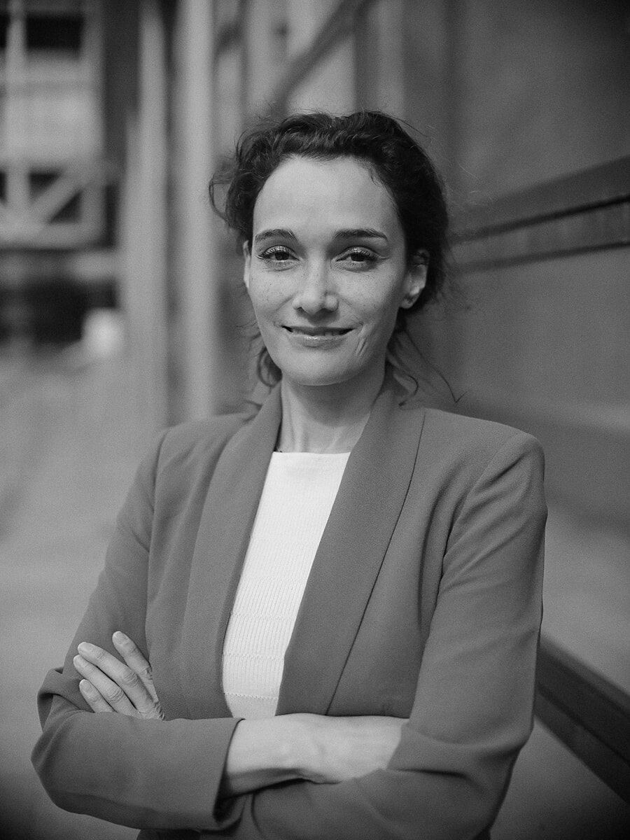 Woman with her hair up and arms folded in a light blazer, an industrial structure blurred behind, a business portrait in Amsterdam