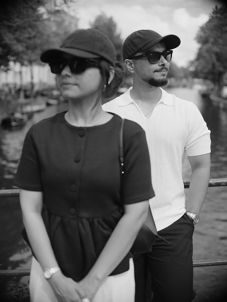 Two people in caps and sunglasses by an Amsterdam canal railing, a black-and-white couple portrait