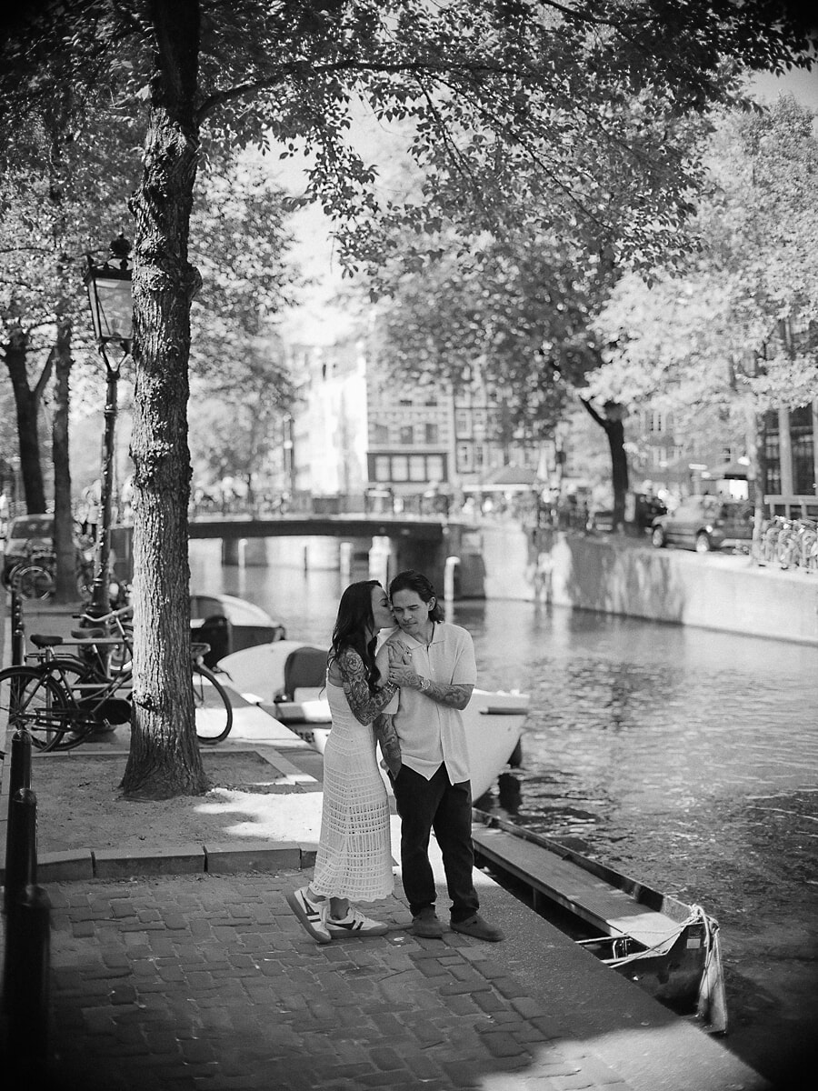 Black-and-white frame of a couple on a cobbled canal quay in Amsterdam, she kisses his cheek beside the moored boats