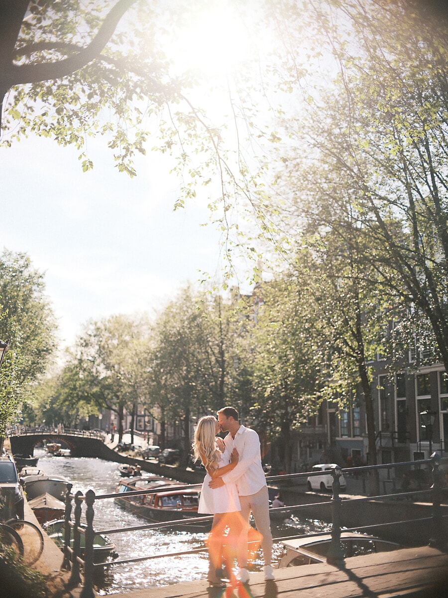 Couple in white kissing at a canal railing under big sunlit trees, a warm lens flare across their engagement shoot in Amsterdam
