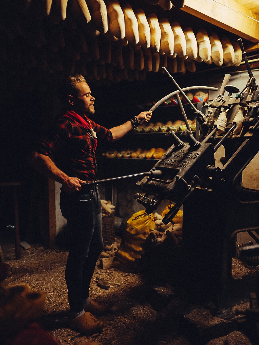 Craftsman working a cast-iron clog-turning machine, rows of wooden clogs hanging from the beams above him