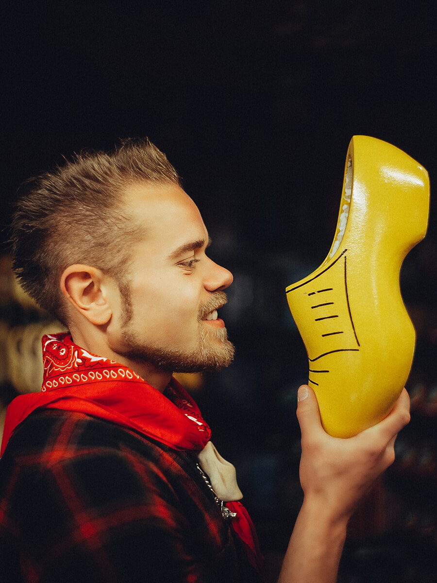 Craftsman in a red bandana holding up a finished yellow clog to inspect it in a dark workshop