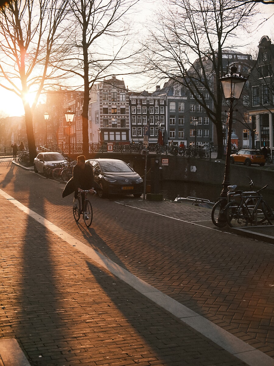 Cyclist riding an Amsterdam canal quay in low winter sun, bare trees casting long shadows, a wide frame from the portrait photoshoot