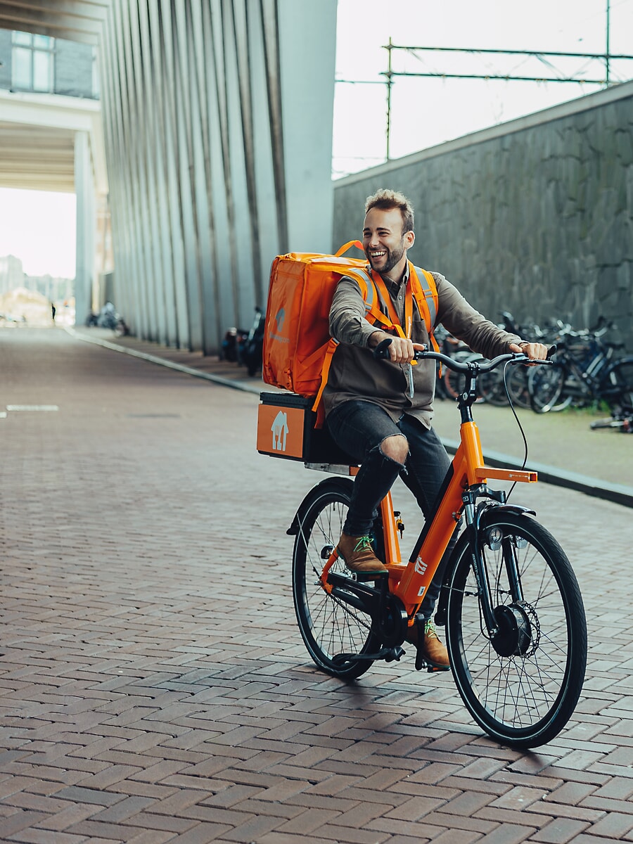 Delivery rider laughing on an orange e-bike with an insulated backpack, on a brick street below a modern building in Amsterdam
