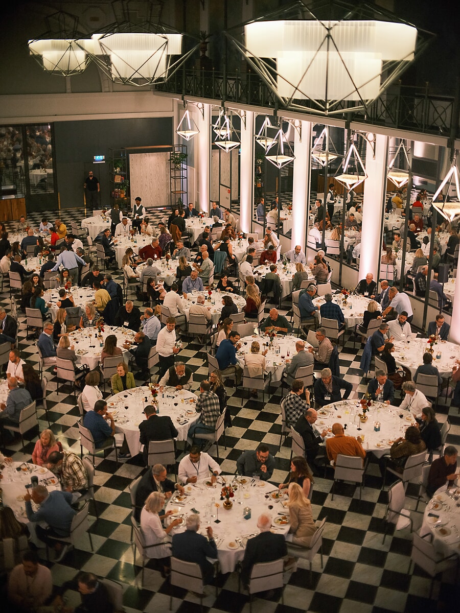 Dining hall full of guests seen from above, round tables on a chequerboard floor beneath geometric lights, event photography in Amsterdam