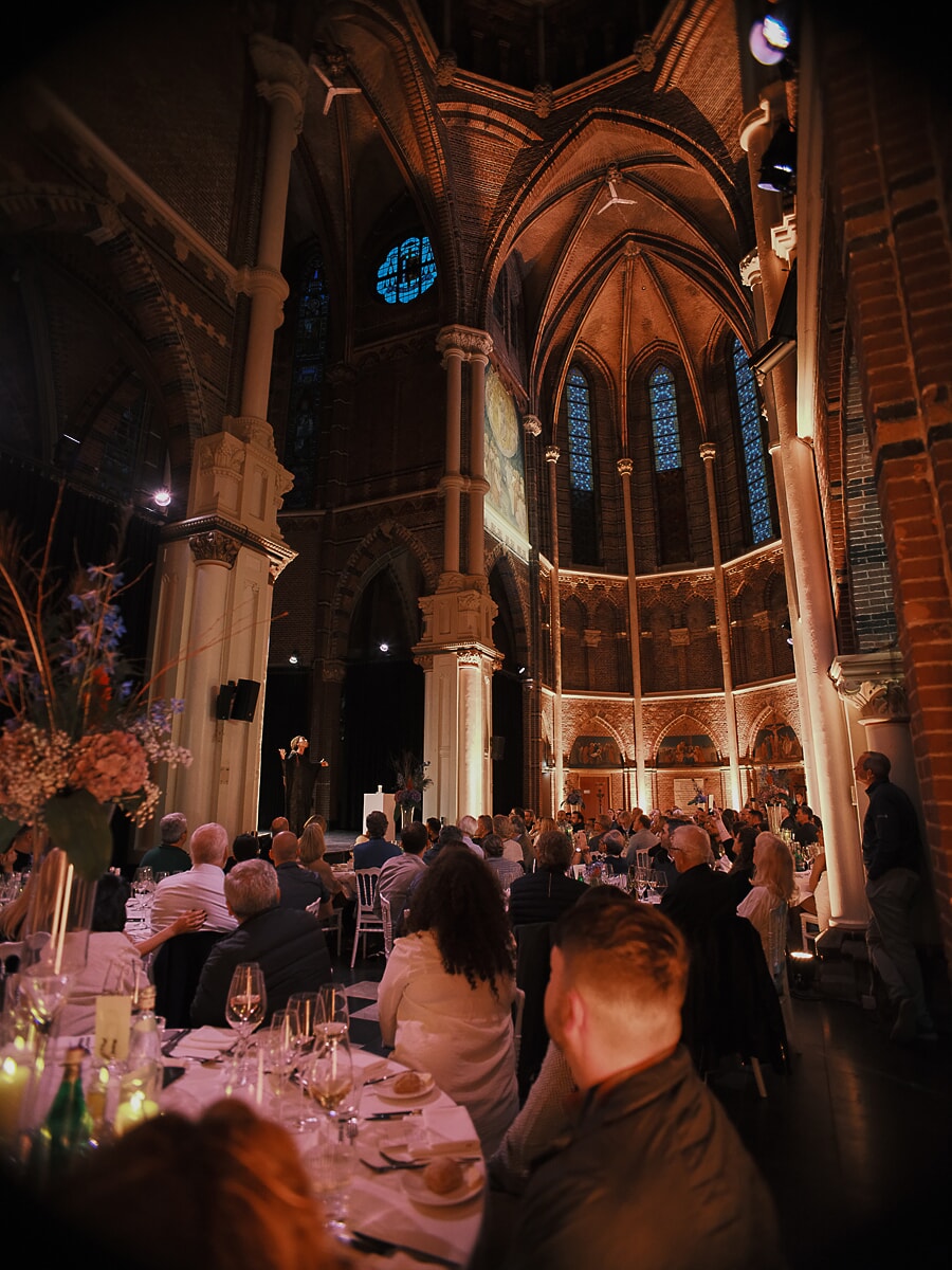 Dinner in a gothic church nave, a speaker in the distance below brick vaults and stained glass, a business event in Amsterdam