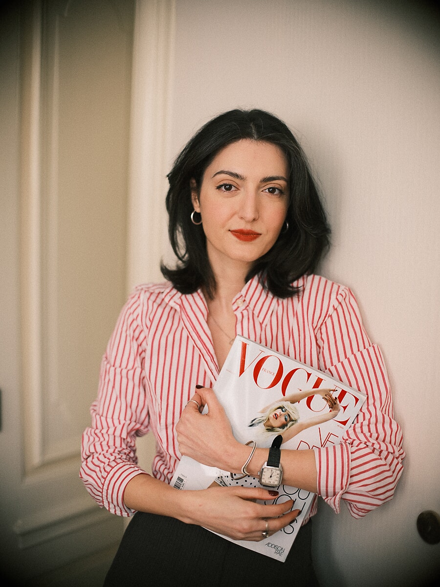 Editor in a red-striped shirt holding a stack of fashion magazines against a panelled cream wall, a branding photoshoot for an Amsterdam business