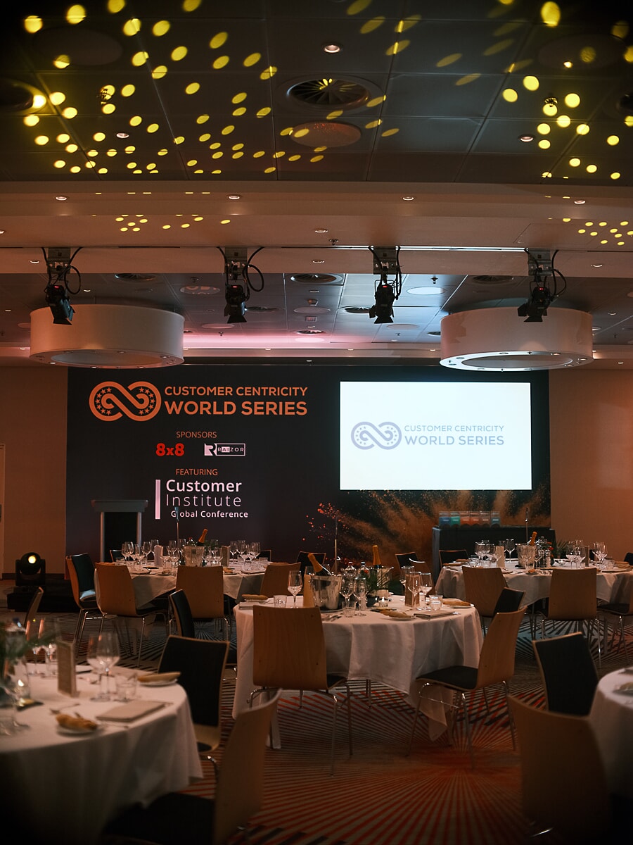 Conference dinner room standing empty, round tables and a lit backdrop under patterned ceiling lights, an Amsterdam business venue
