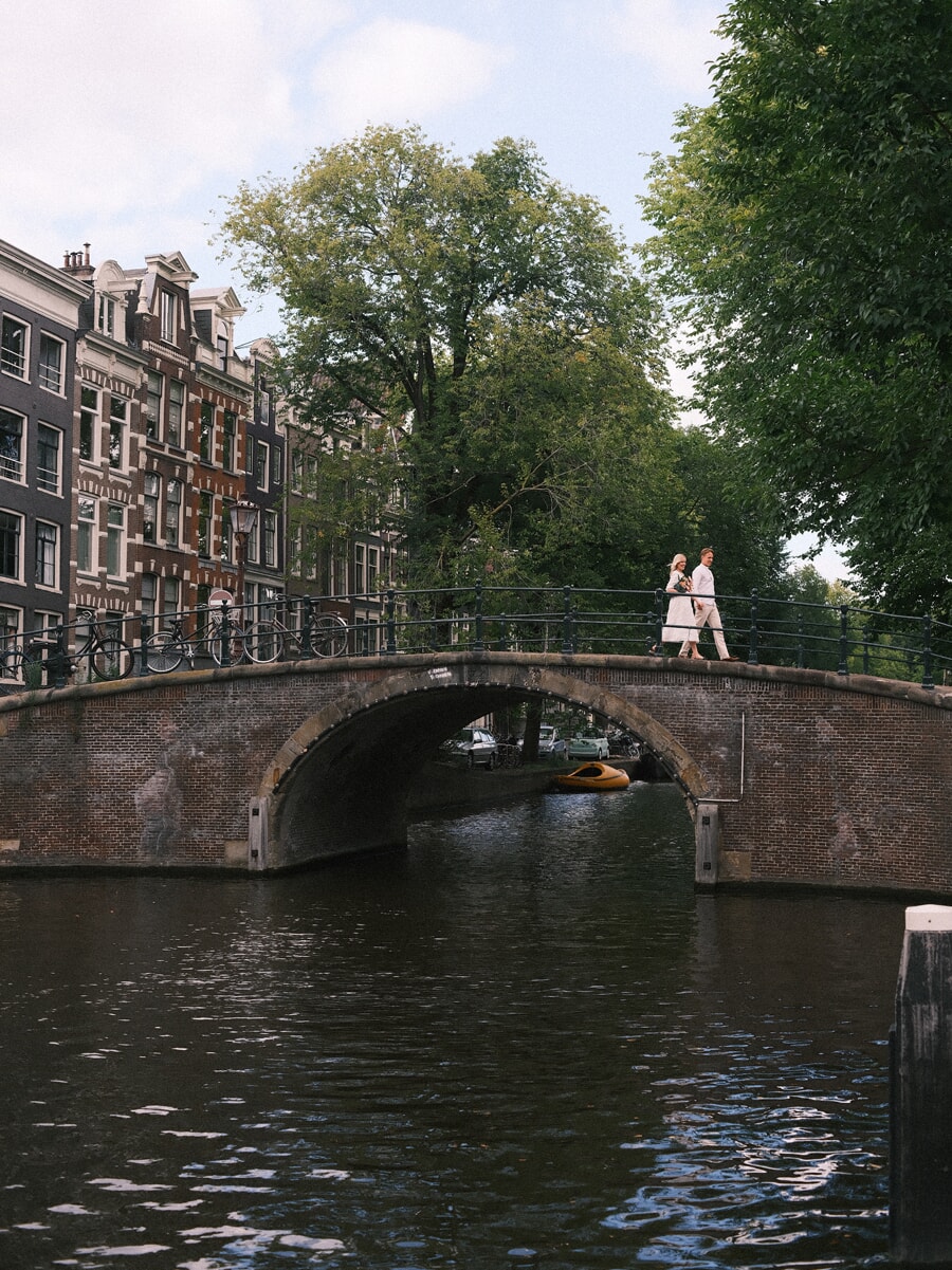 Engaged couple crossing an arched Amsterdam canal bridge, seen wide from the water below