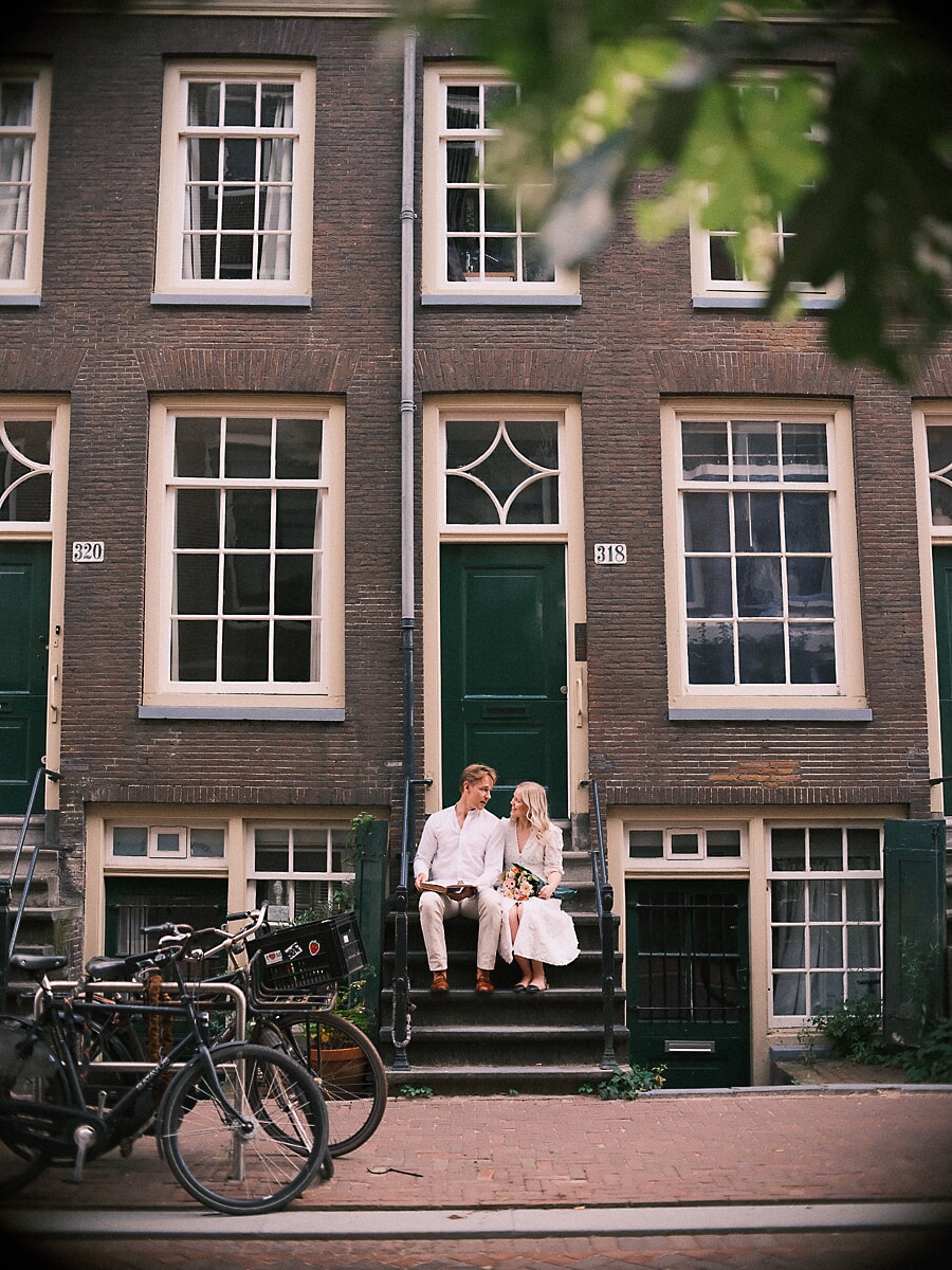 Engaged couple sitting on the stoop of an Amsterdam canal house during their engagement shoot, bicycles along the street