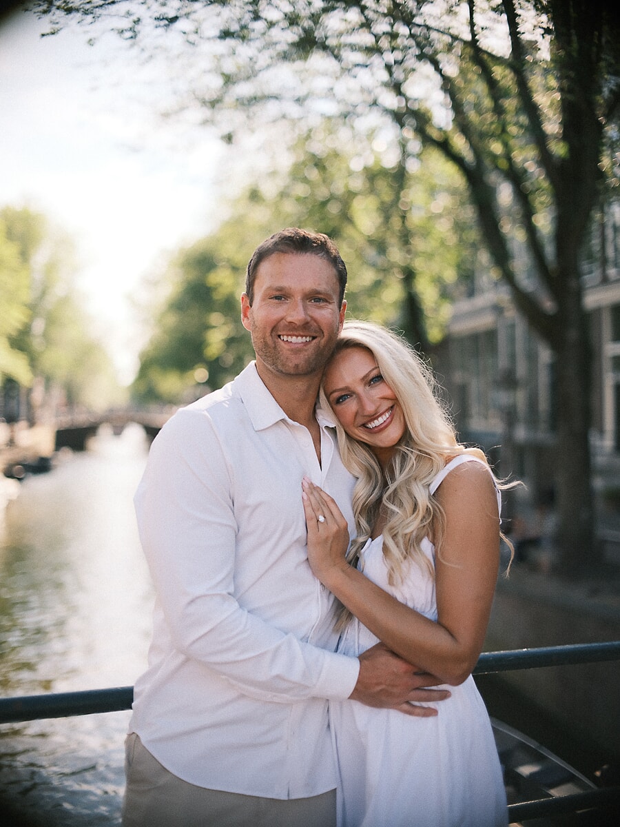 Engaged couple in white smiling by the water, her ring hand resting on his chest, swirly bokeh in the trees behind them