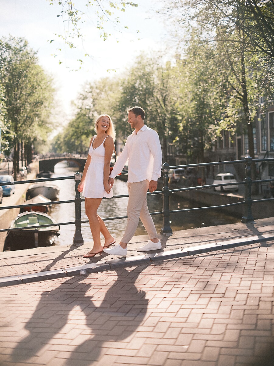 Newly engaged couple walking hand in hand along a sunlit canal quay in Amsterdam, she looks back over her shoulder laughing