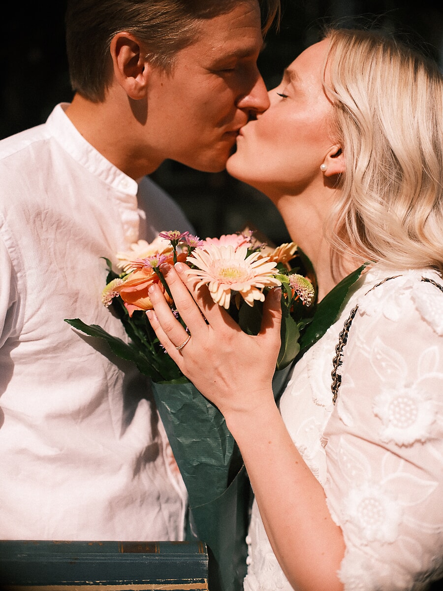 Engaged couple kissing over a bouquet of gerberas, her engagement ring showing against the stems during a pre-wedding session