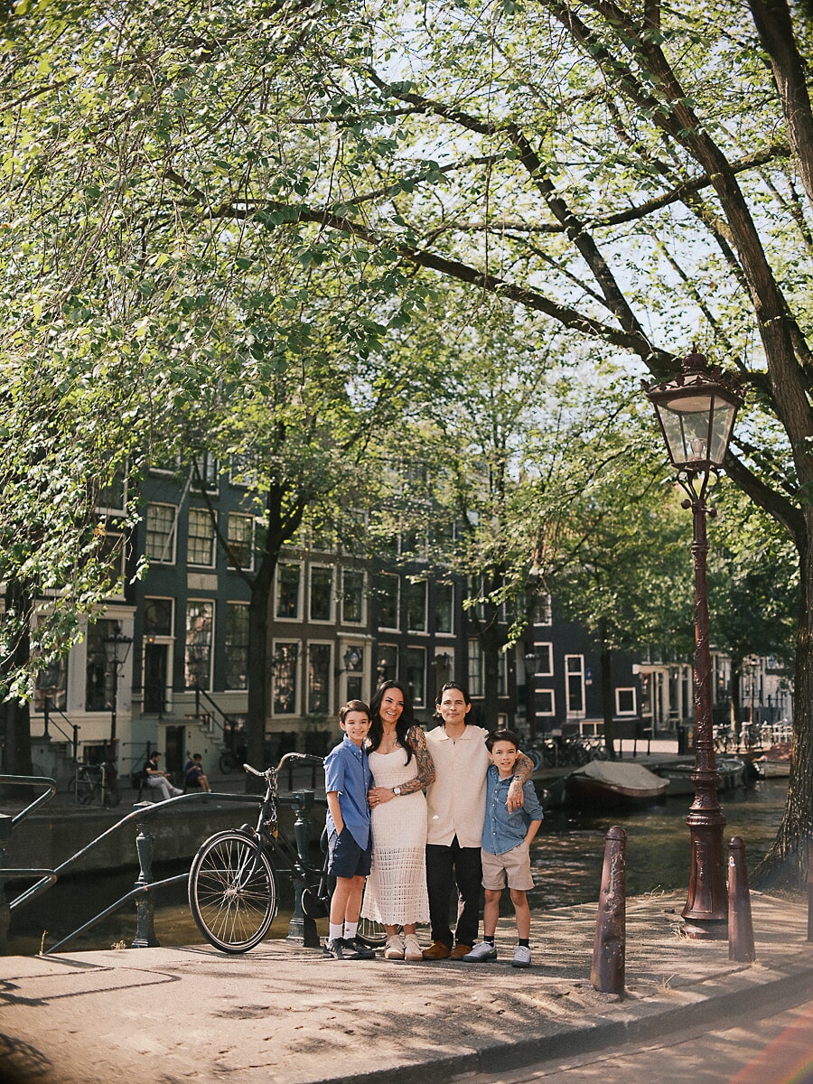 Parents and their two sons arm in arm on a sunlit canal quay in Amsterdam, an ornate lamppost and tall gabled houses behind
